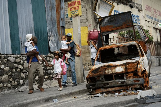 海地街头一辆烧毁的汽车旁，居民牵着孩子在破败环境中行走的场景