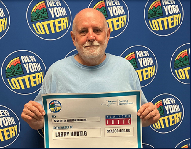 纽约彩票强力球 1700 万美元大奖得主拉里·哈蒂格（Larry Hartig）在领奖大厅拍照留念。他手持一張寫有 $17,000,000.00 金額的巨大支票，證實這位纽约州彩民在時隔三月後現身兌獎的新闻事件。