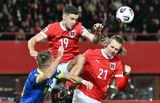 奧地利男足(紅色 19 號和 21 號球員)與波黑男足(藍色球衣)球員在空中激烈爭搶頭球的瞬間。這張照片捕捉了世預賽小組賽 H 組中,奧地利為爭奪世界杯直通名額而進行的身體對抗,該場比賽以 1-1 結束。