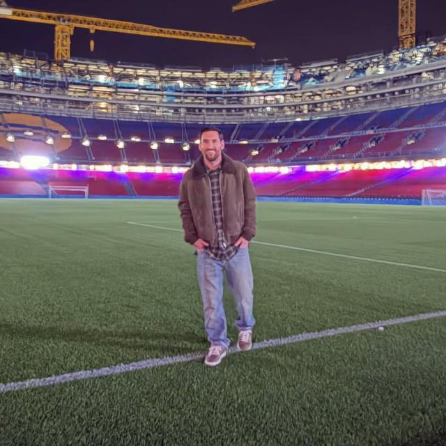 梅西夜间站在施工中的诺坎普草坪上合影，球场灯光与看台起重机清晰可见。