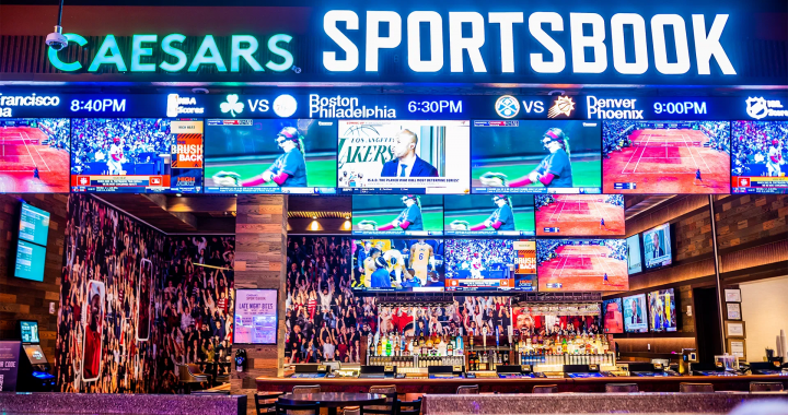 一间现代化的体育博彩区内部，巨大的屏幕上显示着“CAESARS SPORTSBOOK”标志和多场体育比赛的直播画面，吧台前摆放着桌椅供顾客观赛。