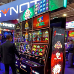 SYNOT集团在2025年BEGE展会上的展位照片，展示了多台带有红色边框的最新游戏机柜，屏幕上播放着老虎机游戏画面，几位参观者正在操作台前进行体验。