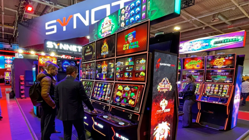 SYNOT集团在2025年BEGE展会上的展位照片，展示了多台带有红色边框的最新游戏机柜，屏幕上播放着老虎机游戏画面，几位参观者正在操作台前进行体验。