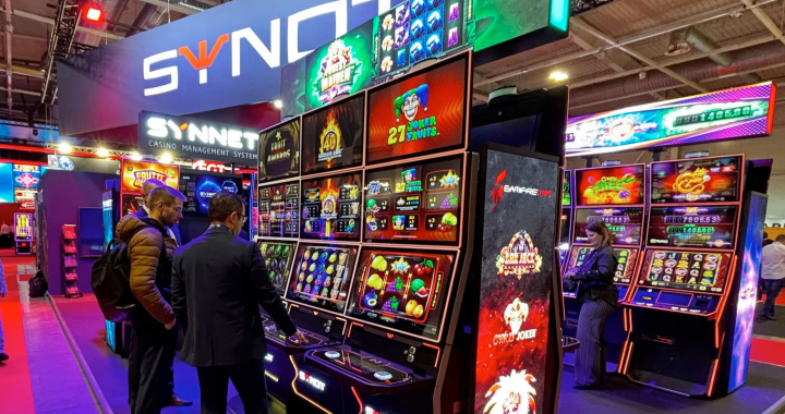 SYNOT集团在2025年BEGE展会上的展位照片，展示了多台带有红色边框的最新游戏机柜，屏幕上播放着老虎机游戏画面，几位参观者正在操作台前进行体验。