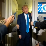 美国总统唐纳德·特朗普身穿深蓝色西装，戴着亮蓝色领带，站在空军一号机舱内，被一群记者和摄像机包围，正在对媒体发表讲话。