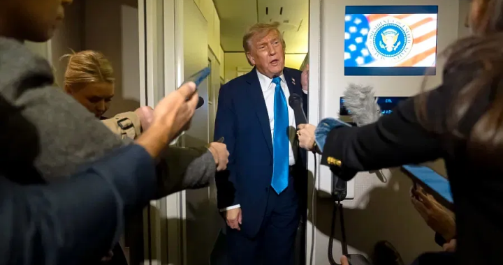 美国总统唐纳德·特朗普身穿深蓝色西装，戴着亮蓝色领带，站在空军一号机舱内，被一群记者和摄像机包围，正在对媒体发表讲话。