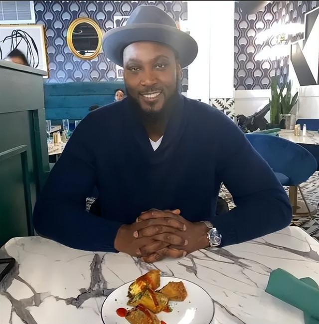 夸梅·布朗坐在餐厅微笑出镜，代表其播客节目《Bust Life》的主持人形象。