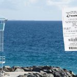 男子在澳大利亞珀斯海灘或海岸邊，手持一張強力球（Powerball）彩票，背景為救生塔和藍色大海