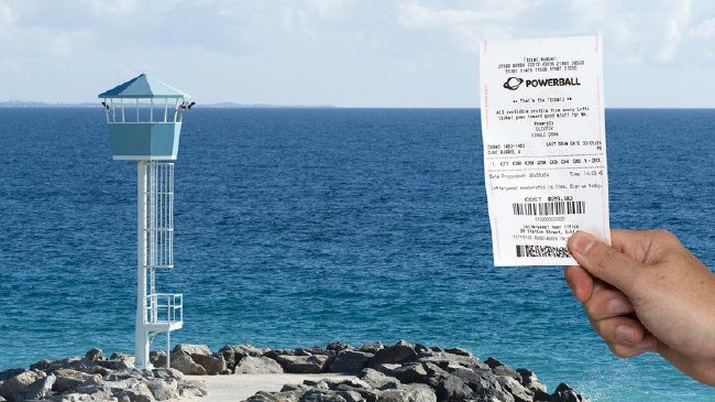 男子在澳大利亞珀斯海灘或海岸邊，手持一張強力球（Powerball）彩票，背景為救生塔和藍色大海
