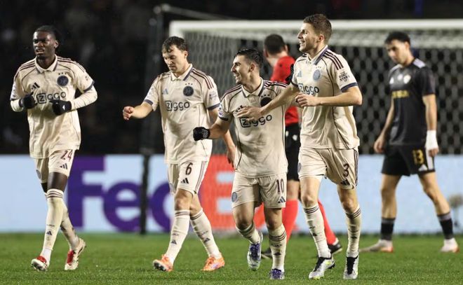 身穿浅色球衣的阿贾克斯足球运动员在球场上奔跑庆祝,其中一名球员(背号10)面露笑容。