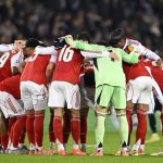 阿森納球員（紅白球衣）在足球場上進行賽前或中場的圓陣喊話，門將穿著螢光綠色球衣，體現團隊士氣。