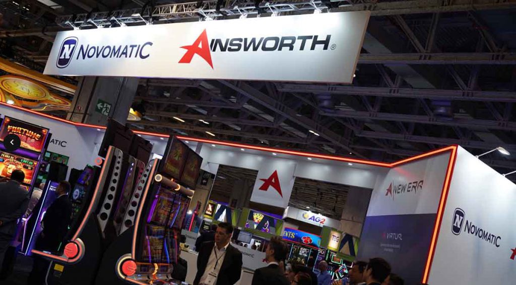 会展现场悬挂Novomatic与Ainsworth品牌招牌，下方陈列多台老虎机设备