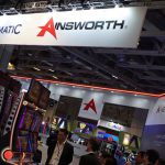 会展现场悬挂Novomatic与Ainsworth品牌招牌，下方陈列多台老虎机设备