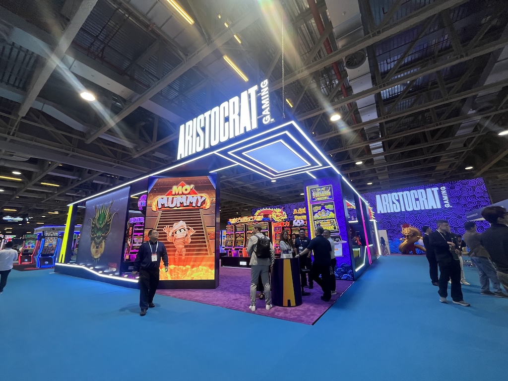 展会现场Aristocrat Gaming展位，参观者在多台电子游戏机前交流，展位悬挂品牌标识灯箱