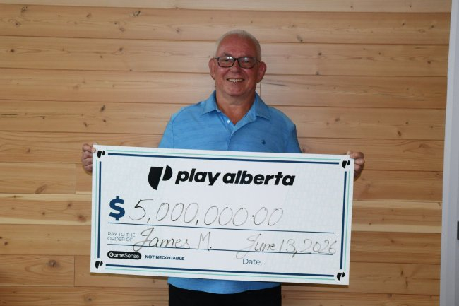 身穿蓝色衬衫的老年男子手持写有五百万美元字样的Play Alberta巨型支票,站在木纹墙面前